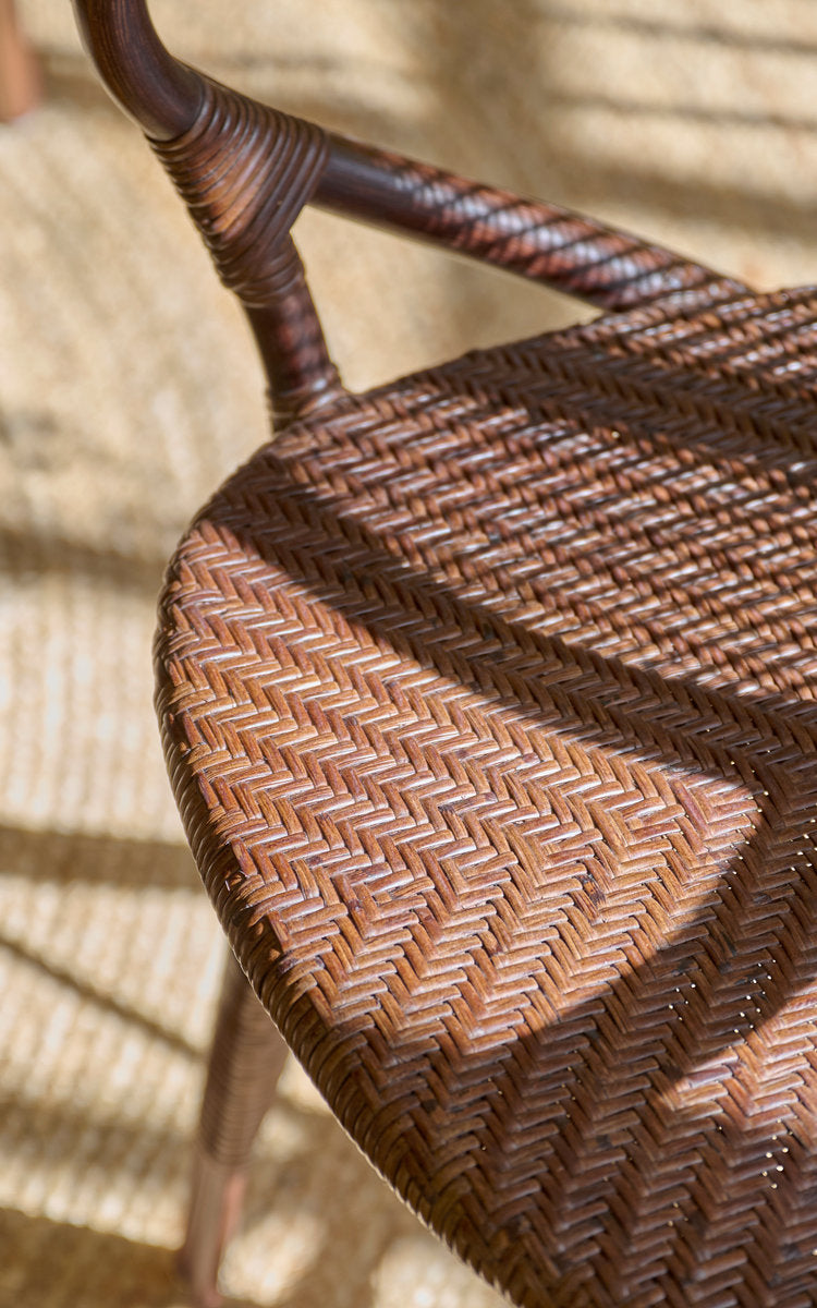 Dies ist ein Nahaufnahmebild von Rossini Esszimmer-Armlehnstuhl in brown rattan. Sitz im Fischgrätgeflecht und umwickelte Armlehne im warmen Sonnenlicht.
