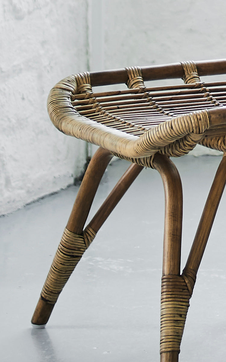 Dies ist ein Nahaufnahmebild von Monet Footstool i antique rattan. Detail der flachen geflochtenen Sitzfläche und der gespreizten Rattanbeine.