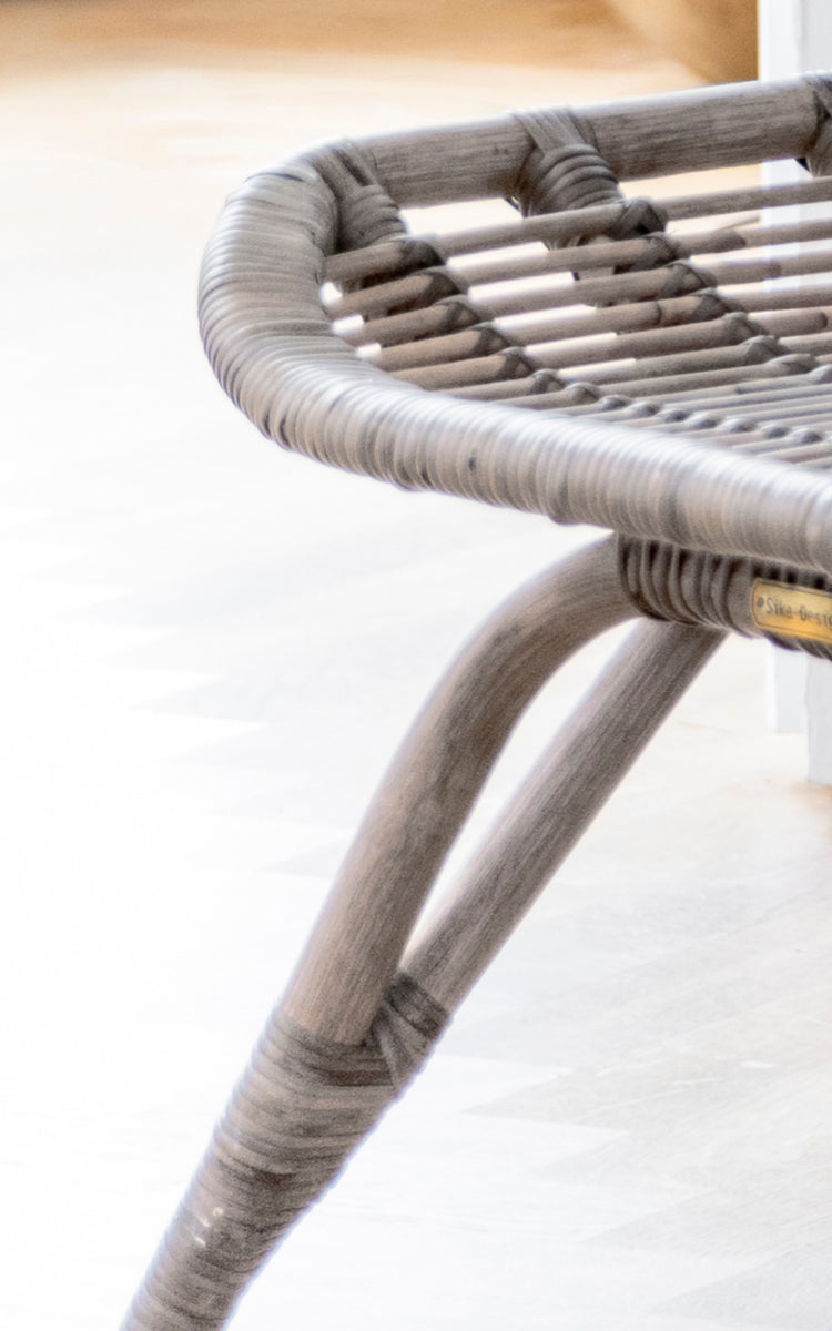 Dies ist ein Nahaufnahmebild von Monet Footstool i taupe rattan. Detail der geflochtenen Sitzfläche und des gebogenen Beingestells in weichem Taupe-Finish.
