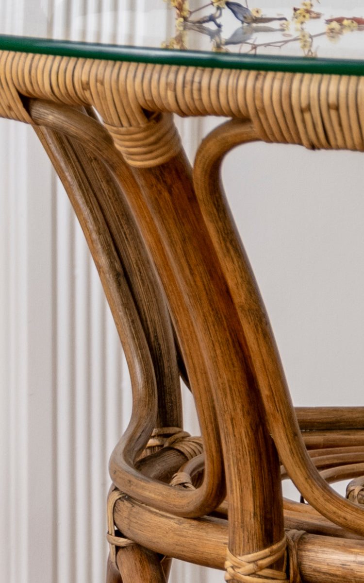 Dies ist ein Nahaufnahmebild von Tony Café Table in antique rattan. Detail der Glasplattenkante, des gebogenen Rattanrahmens und umwickelten Verbindungen.