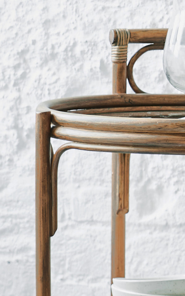 Dies ist ein Nahaufnahmebild von Romeo Servierwagen in antique rattan. Detail des runden Rattan-Servierwagen-Rahmens vor einer weißen Backsteinwand.