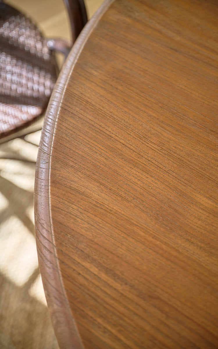 Dies ist ein Nahaufnahmebild von Michel Teak-Esstisch Ø120 cm in teak indoor. Kante einer runden Teakplatte mit Rattanstuhl dahinter.
