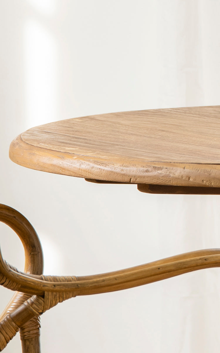 Dies ist ein Nahaufnahmebild von Michel Teak Dining Table Ø80 cm in teak indoor. Die runde Tischplattenkante und der geschwungene Rattan-Säulenfuß sind gezeigt.