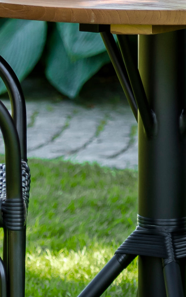 Dies ist ein Nahaufnahmebild von Nicole Cafe Table i black aluminium frame black ArtFibre. Detail der Teak-Platte und des umwickelten Fußes.