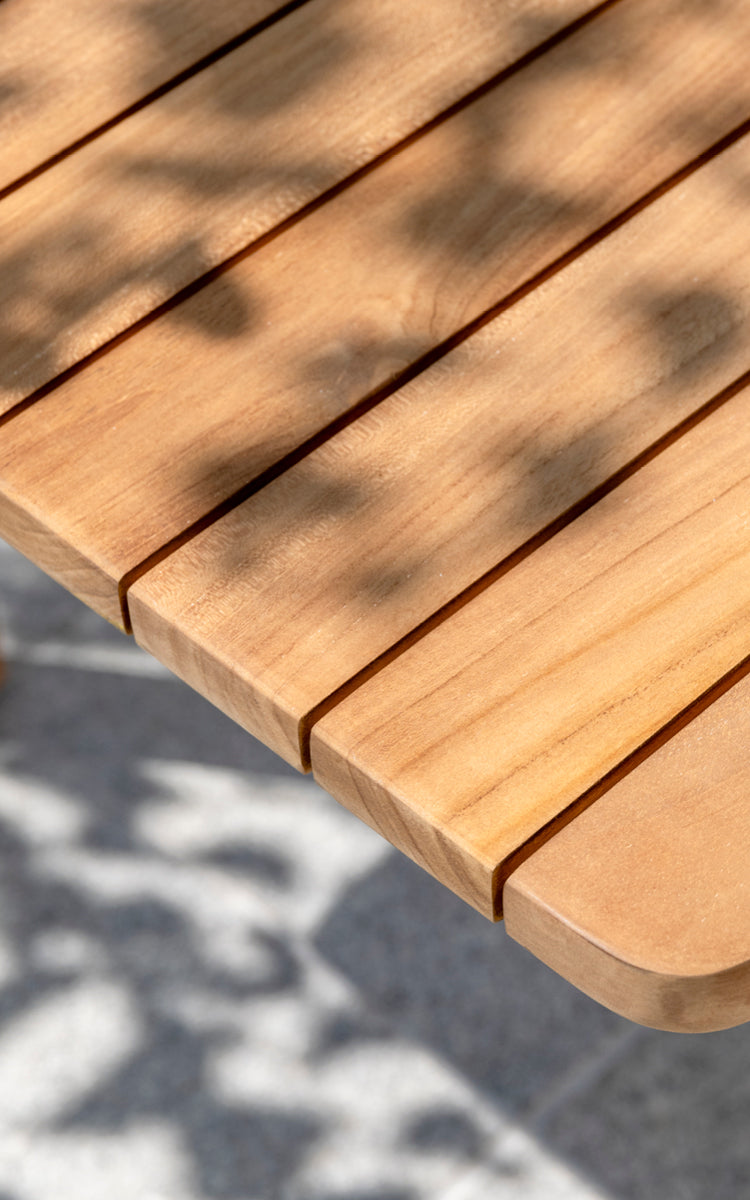 Dies ist ein Nahaufnahmebild von Tobias Exterior Teak Table 205x91 cm in teak outdoor. Detail der Teakbohlen-Tischplattenkante und des gesprenkelten Sonnenlichts.