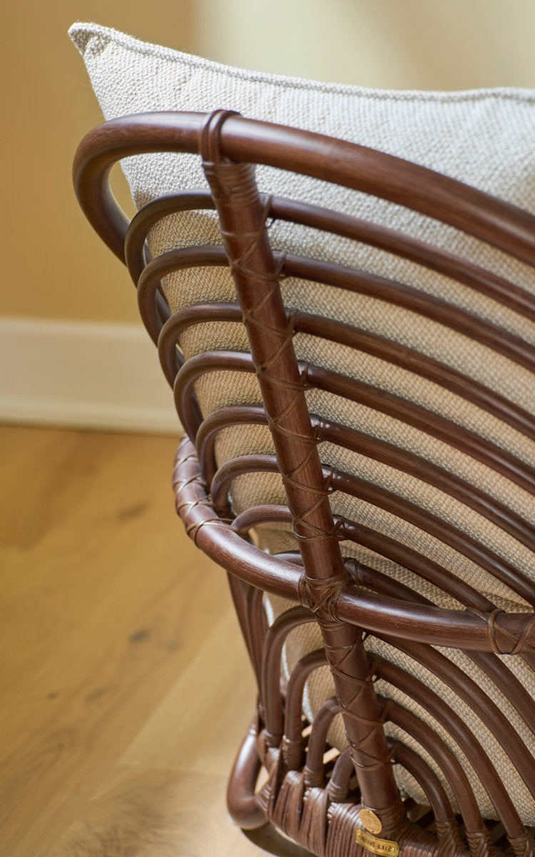 Dies ist ein Nahaufnahmebild von Charlottenborg Lounge-Sessel in brown rattan. Geschwungene Rattanlehne mit Leinenkissen.