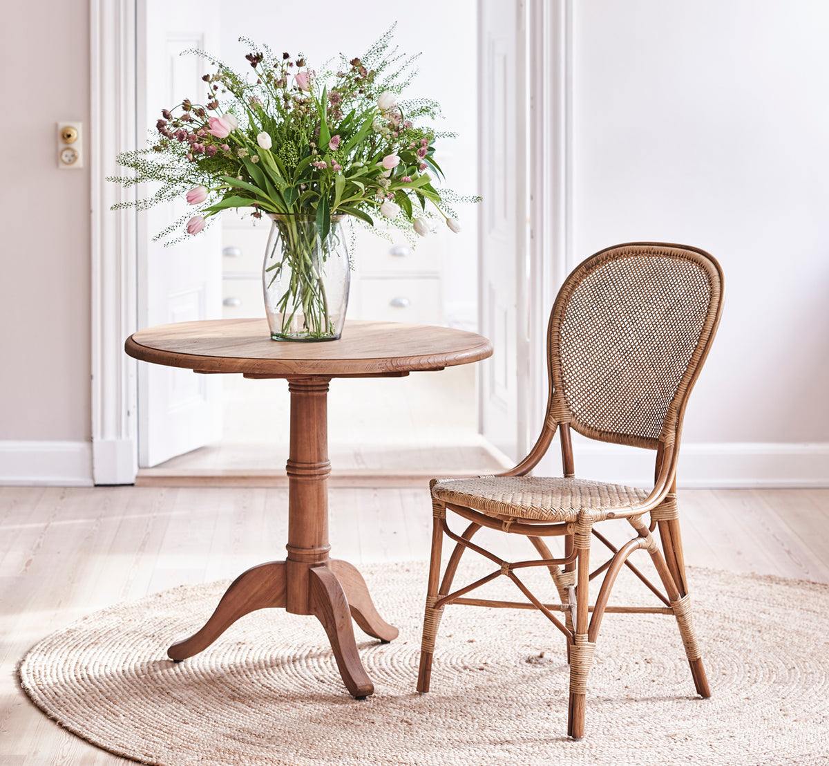 Dies ist ein Lifestyle-Bild von Rossini Dining Chair i antique rattan. Um einen runden Teaktisch auf einem Juteteppich in einem hellen Loft angeordnet.