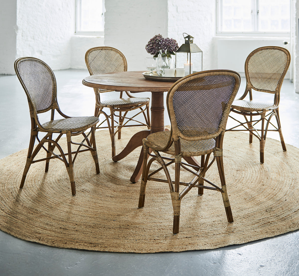 Dies ist ein Lifestyle-Bild von Rossini Dining Chair i antique rattan. Vier Stühle um einen runden Teaktisch auf einem Juteteppich in einem weißen Loft.