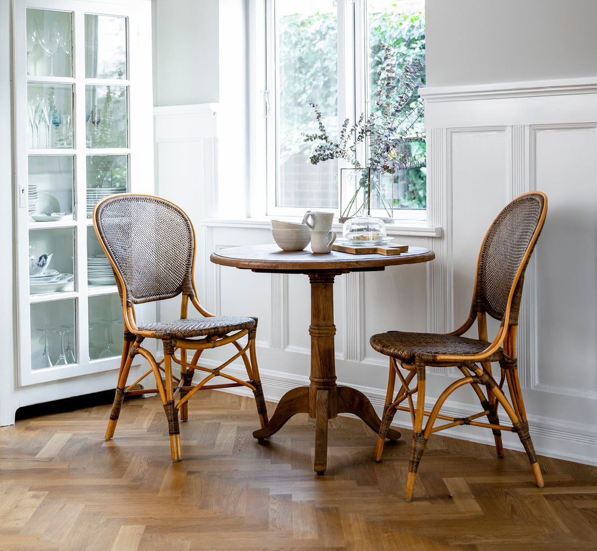 Dies ist ein Lifestyle-Bild von Rossini Dining Chair i cherry rattan. Zwei Stühle an einem kleinen Tisch in einem Innenhof mit Schachbrettfliesen.