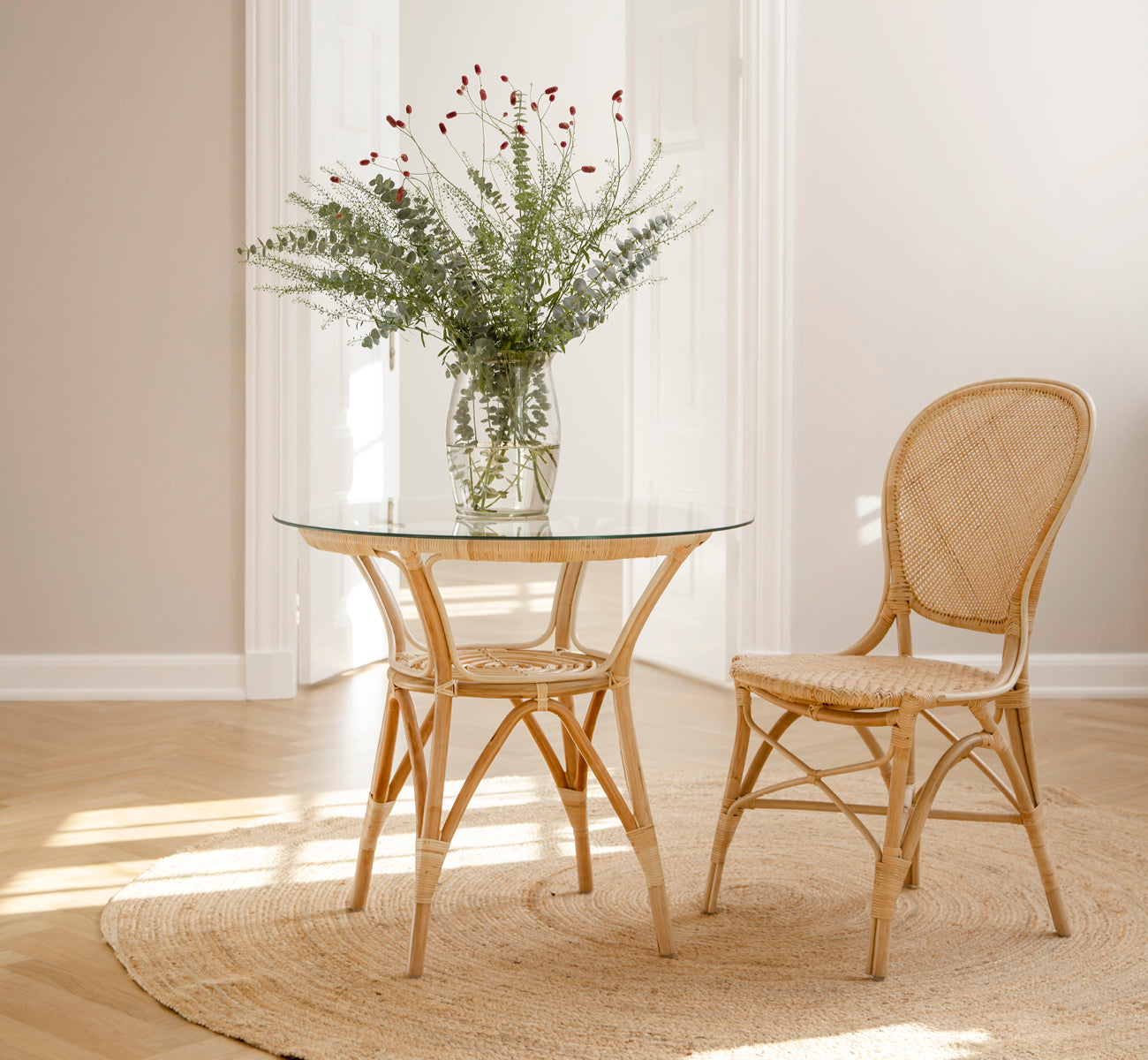 Dies ist ein Lifestyle-Bild von Rossini Esszimmerstuhl in natural rattan. Neben einem Bistrotisch mit Glasplatte auf einem Juteteppich in einem hellen Flur platziert.