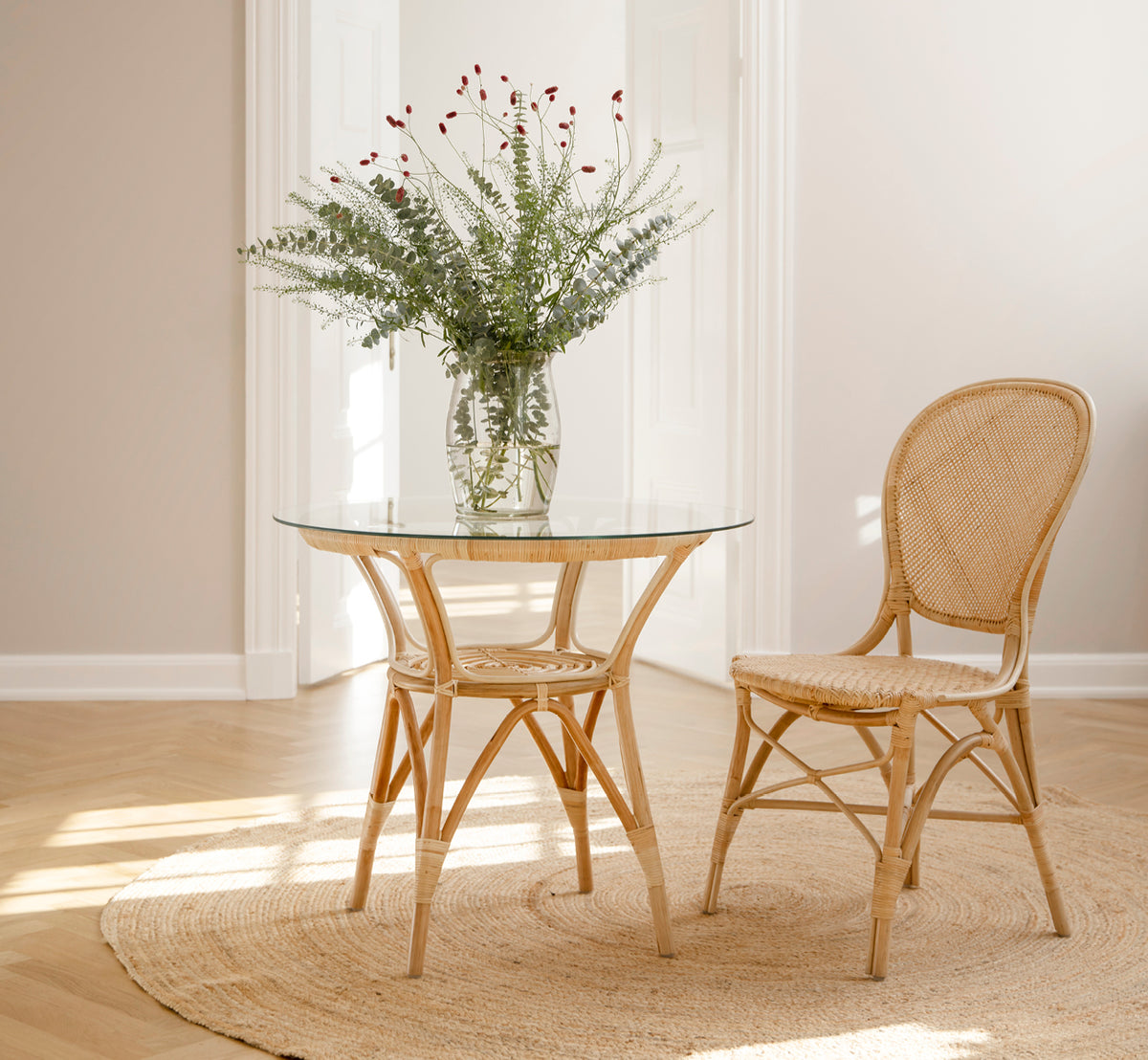 Dies ist ein Lifestyle-Bild von Rossini Esszimmerstuhl in natural rattan. Neben einem Bistrotisch mit Glasplatte auf einem Juteteppich in einem hellen Flur platziert.