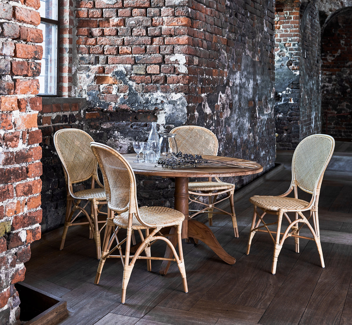 Dies ist ein Lifestyle-Bild von Rossini Esszimmerstuhl in natural rattan. Neben einem Bistrotisch mit Glasplatte auf einem Juteteppich in einem hellen Flur platziert.