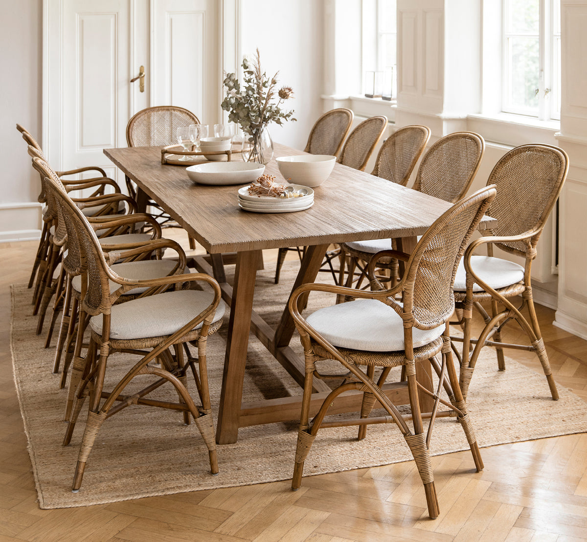 Dies ist ein Lifestyle-Bild von Rossini Esszimmer-Armlehnstuhl in antique rattan. Um einen langen Teaktisch mit Leinenstoff auf einem runden Juteteppich.