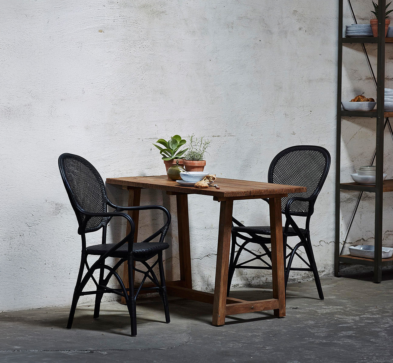 Dies ist ein Lifestyle-Bild von Rossini Esszimmer-Armlehnstuhl in black rattan. Um einen Recyclingholztisch in einem rohen Betonloft angeordnet.