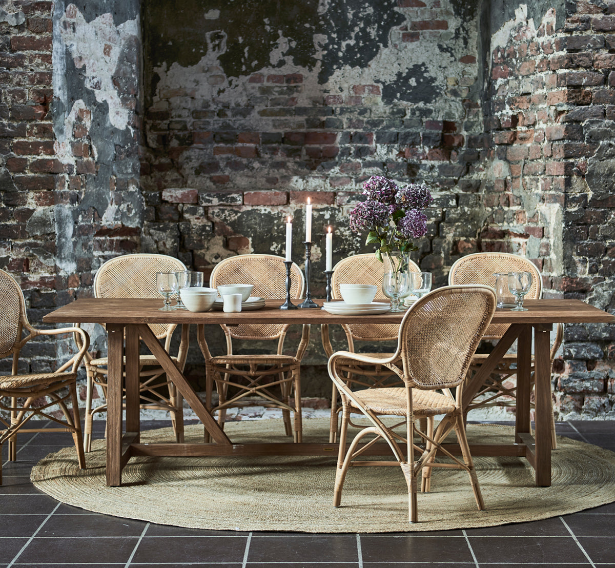 Dies ist ein Lifestyle-Bild von Rossini Esszimmer-Armlehnstuhl in natural rattan. Mit einem runden Glastisch auf einem Juteteppich in einem vertäfelten Raum kombiniert.