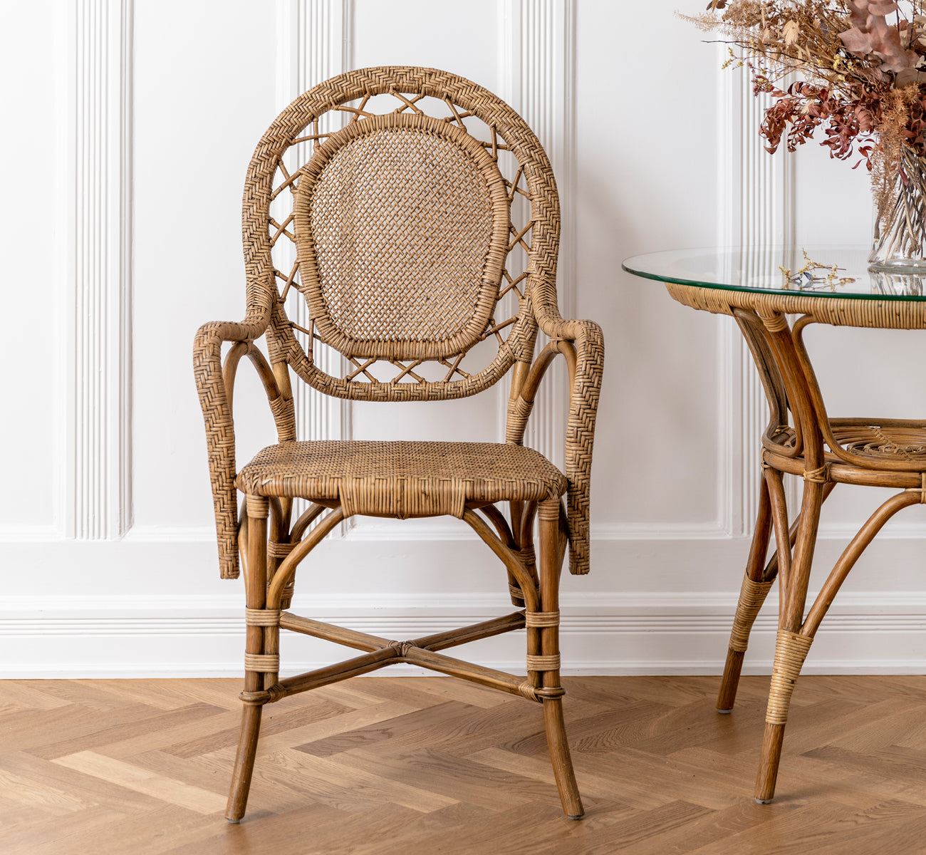 Dies ist ein Lifestyle-Bild von Romantica Esszimmerstuhl in antique rattan. Neben einem Glas-Rattantisch mit Trockenblumen in einem vertäfelten Raum platziert.
