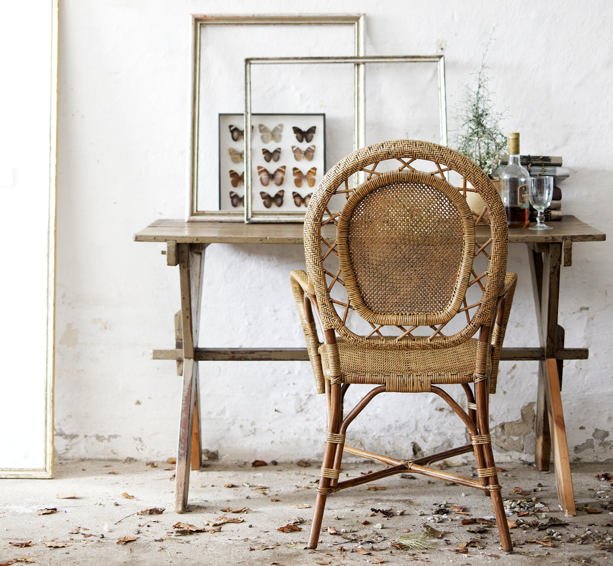 Dies ist ein Lifestyle-Bild von Romantica Esszimmerstuhl in antique rattan. An einem rustikalen Holztisch mit gerahmter Kunst in einem Vintage-Ambiente gestylt.