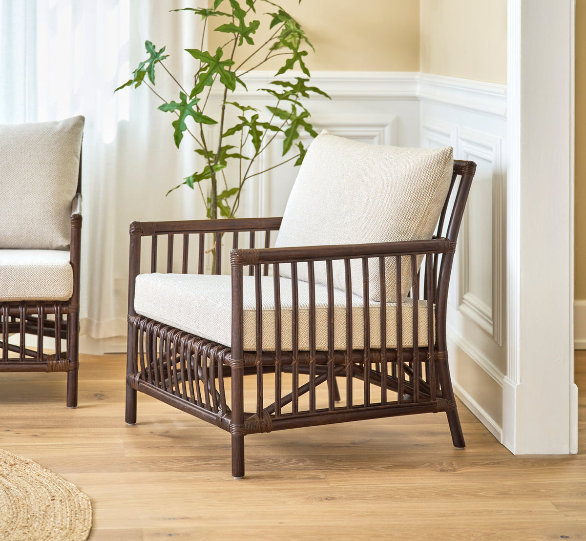 Dies ist ein Lifestyle-Bild von Caroline Lounge-Sessel in brown rattan. Mit Leinenkissen neben weiß vertäfelten Wänden.