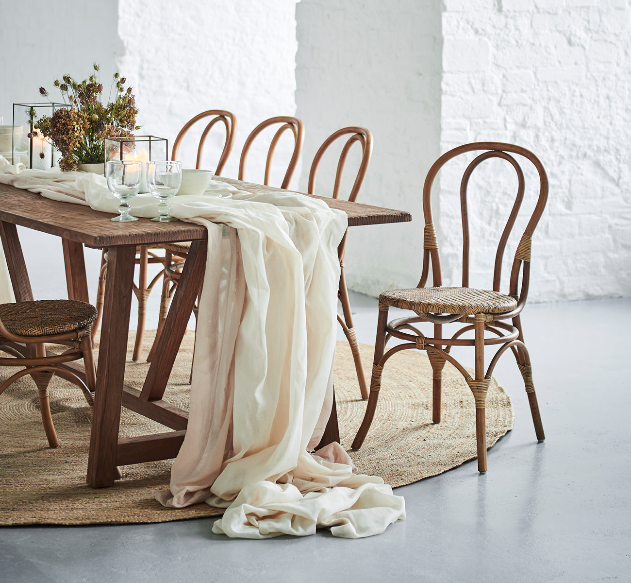 Dies ist ein Lifestyle-Bild von Lulu Esszimmerstuhl in antique rattan. An einem rustikalen Tisch mit Leinenstoff und Kerzen vor einer weißen Backsteinwand.