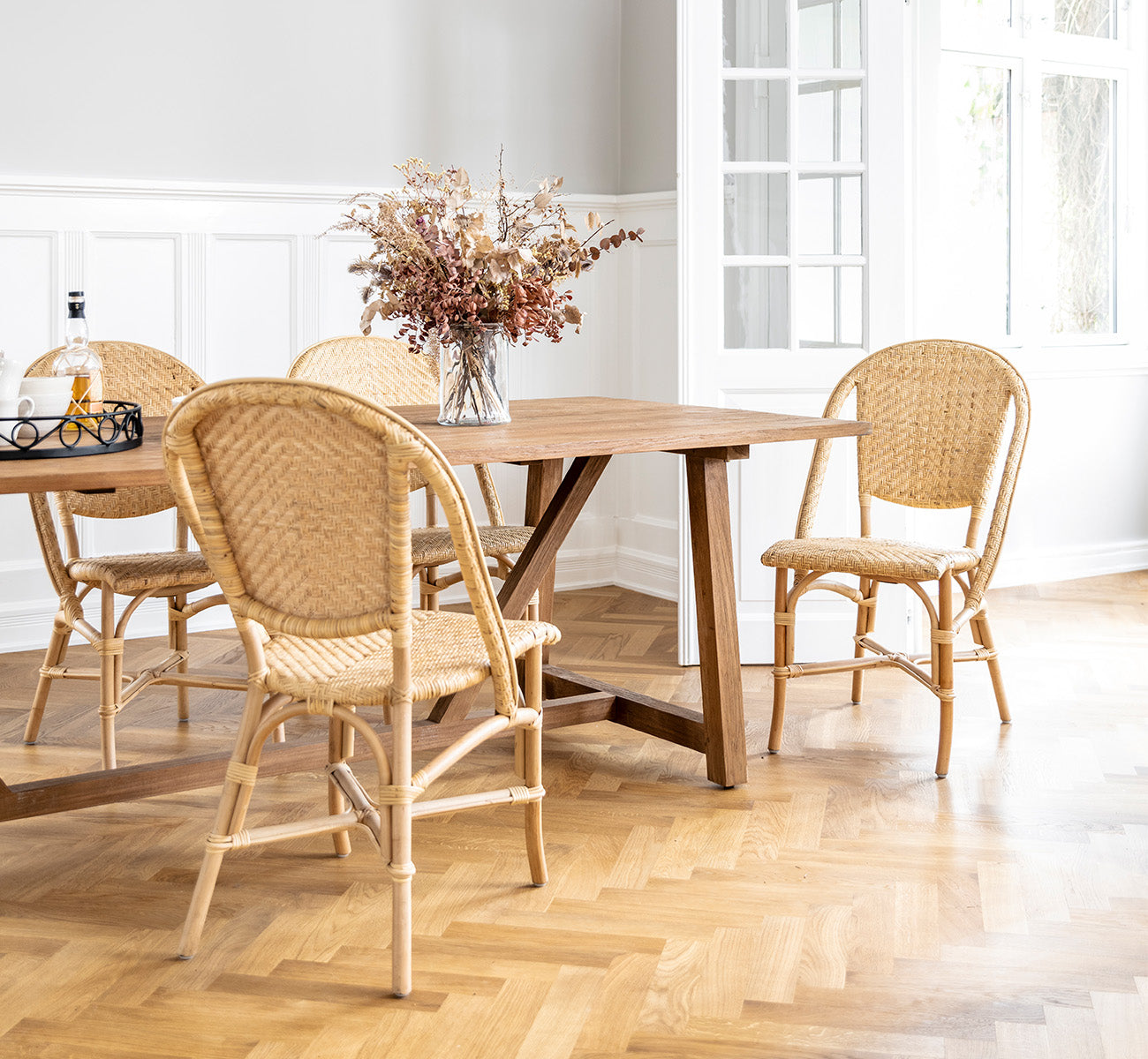 Dies ist ein Lifestyle-Bild von Alanis Esszimmerstuhl in natural rattan, in einem hellen Interieur mit weißen Wandpaneelen und Fischgrätparkett.
