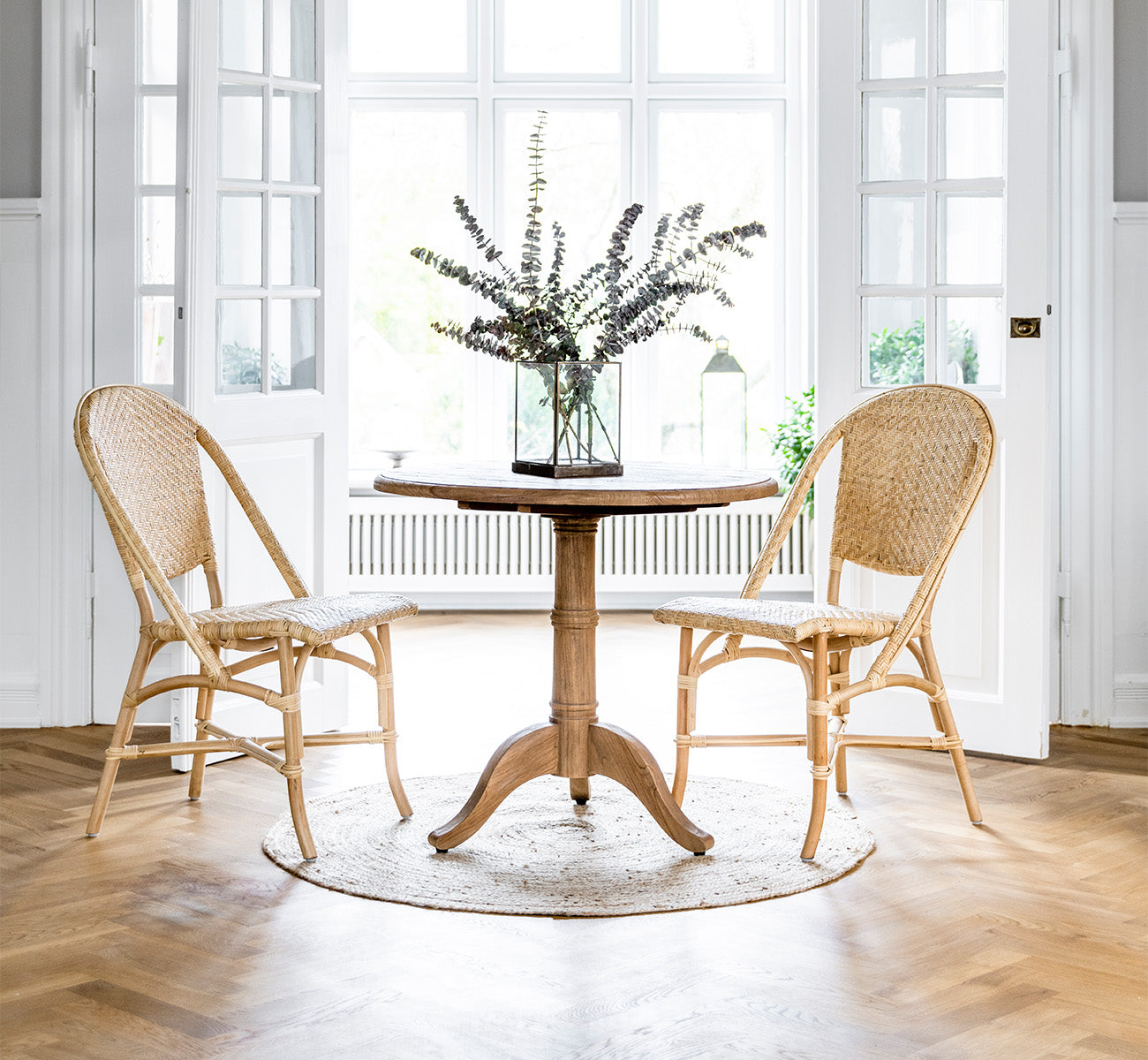 Dies ist ein Lifestyle-Bild von Alanis Esszimmerstuhl in natural rattan, in einem hellen Interieur mit weißen Wandpaneelen und Fischgrätparkett.