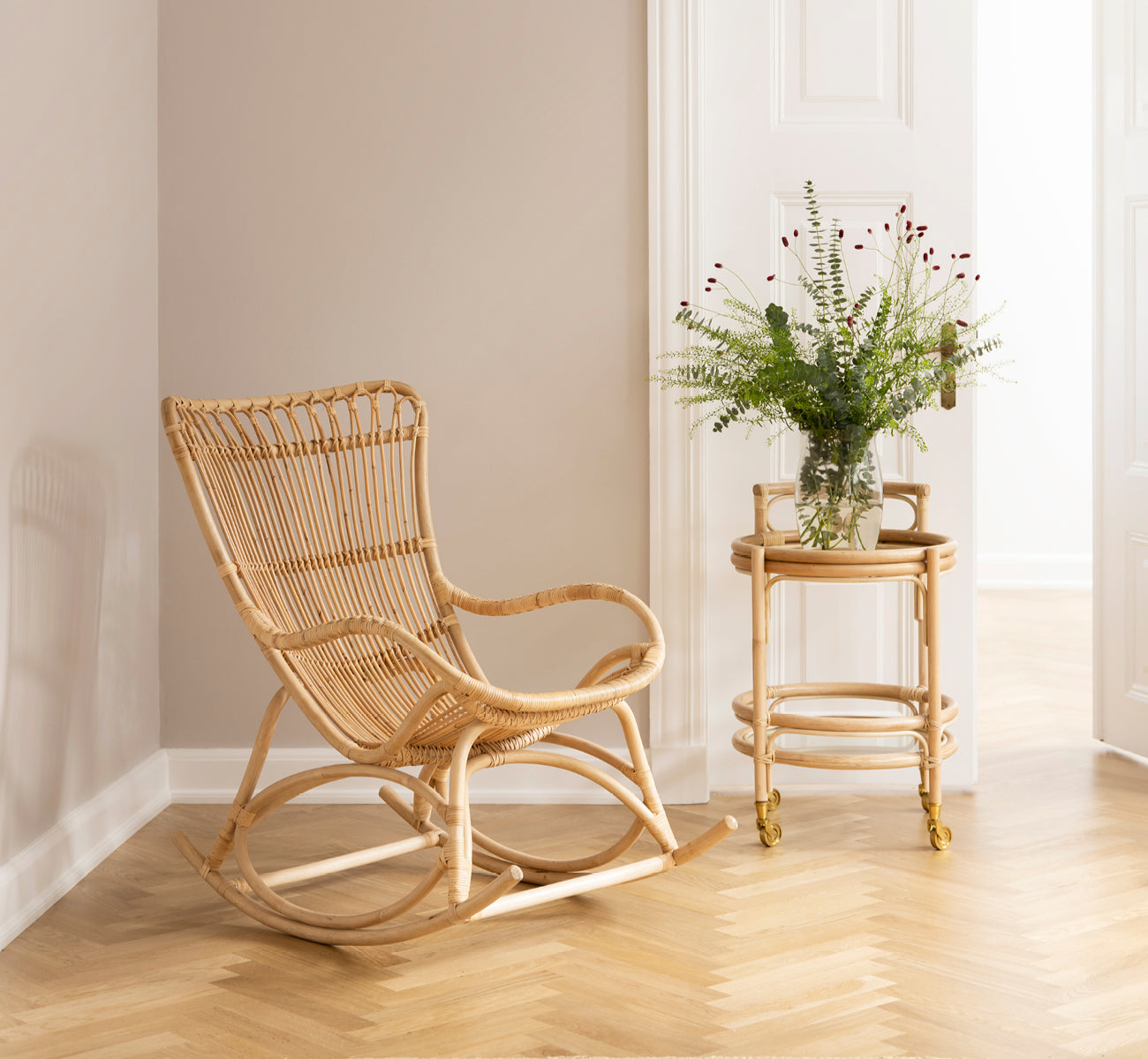 Dies ist ein Lifestyle-Bild von Monet Rocking Chair i natural rattan. In einer gemütlichen Ecke mit Fischgrätboden und Rattan-Servierwagen.
