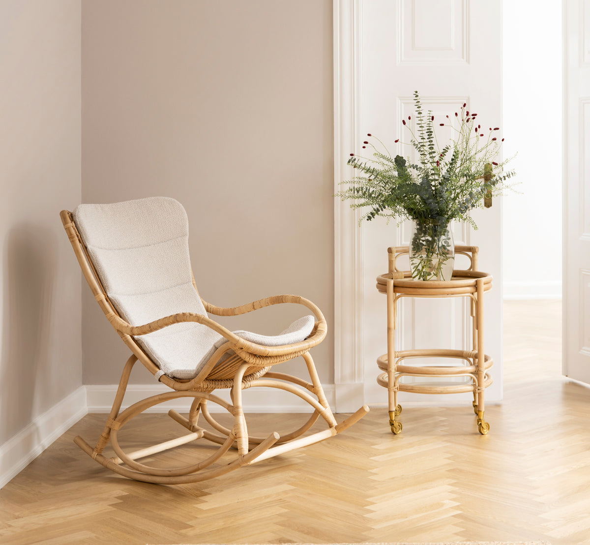 Dies ist ein Lifestyle-Bild von Monet Rocking Chair i natural rattan. In einem Raum mit rosafarbenen Wänden, Fischgrätparkett und Eukalyptus.