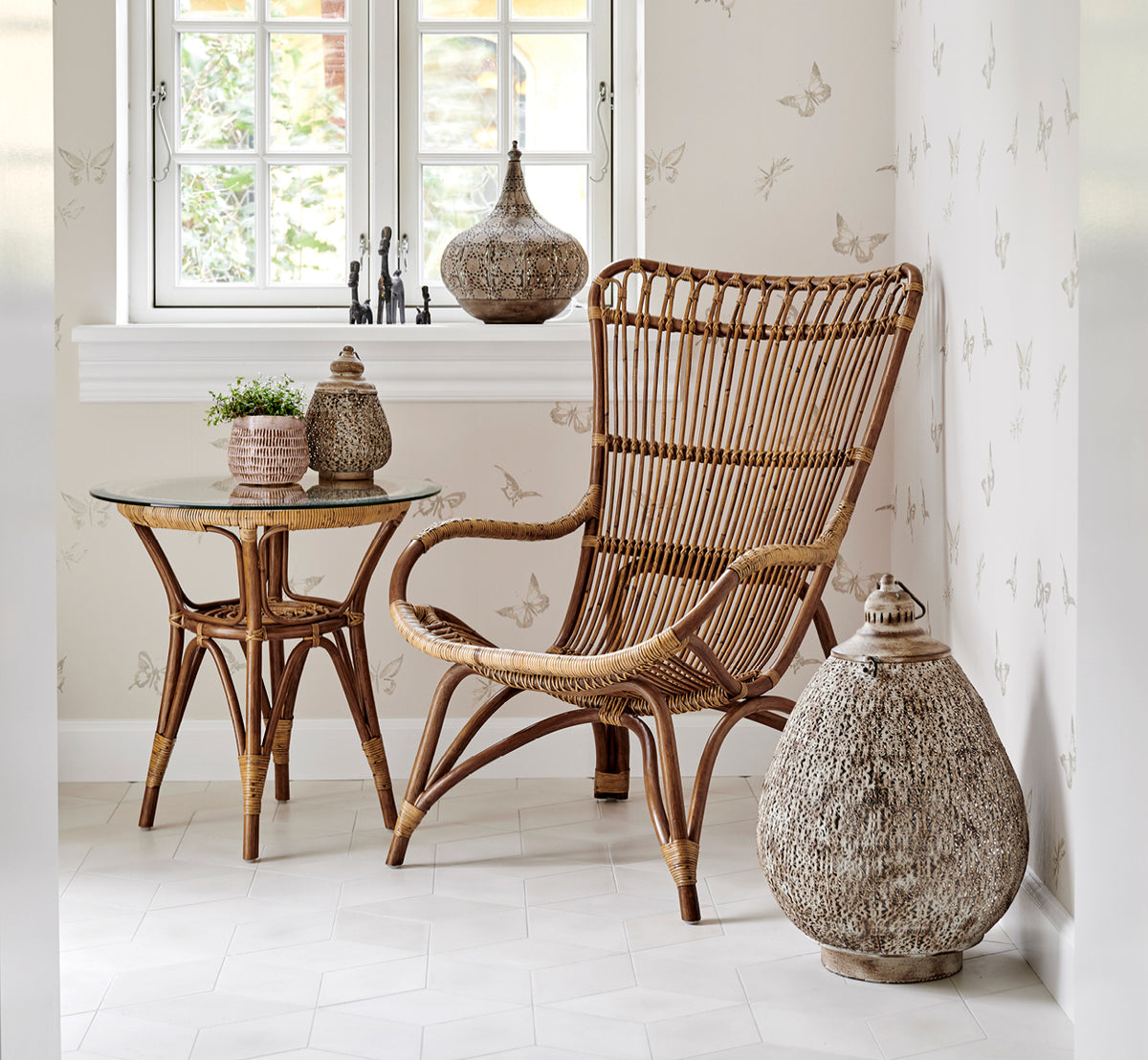 Dies ist ein Lifestyle-Bild von Monet Lounge Chair i antique rattan. Mit Fußhocker in einem rustikalen weißen Raum mit dunklem Marinetextil.