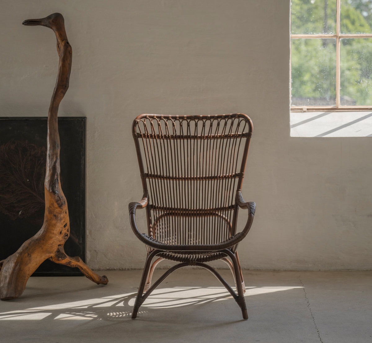 Dies ist ein Lifestyle-Bild von Monet Lounge-Sessel in brown rattan. Neben Treibholz in einem rustikalen Raum mit Betonboden.