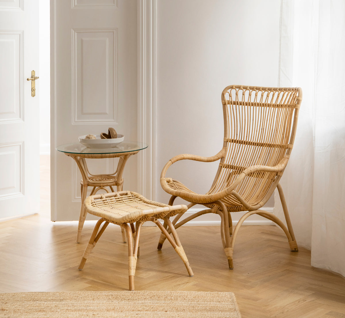 Dies ist ein Lifestyle-Bild von Monet Lounge Chair i natural rattan. Mit Fußhocker und Beistelltisch nahe einer Tür auf Fischgrätboden.