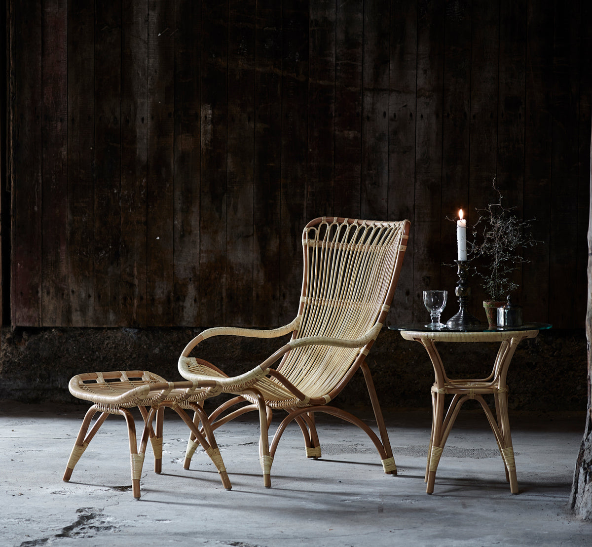 Dies ist ein Lifestyle-Bild von Monet Lounge-Sessel in natural rattan. Mit einem Fußhocker und Beistelltisch vor einer dunklen holzverkleideten Wand kombiniert.
