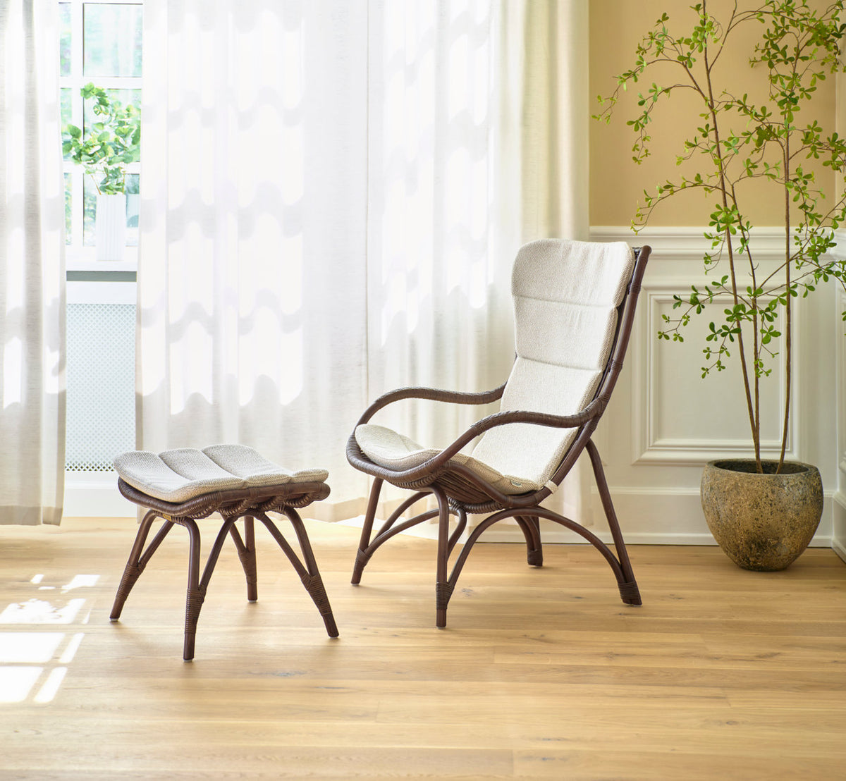 Dies ist ein Lifestyle-Bild von Monet Fußhocker in brown rattan. Mit gepolstertem Lounge-Sessel und Pflanze in einem hellen Raum.