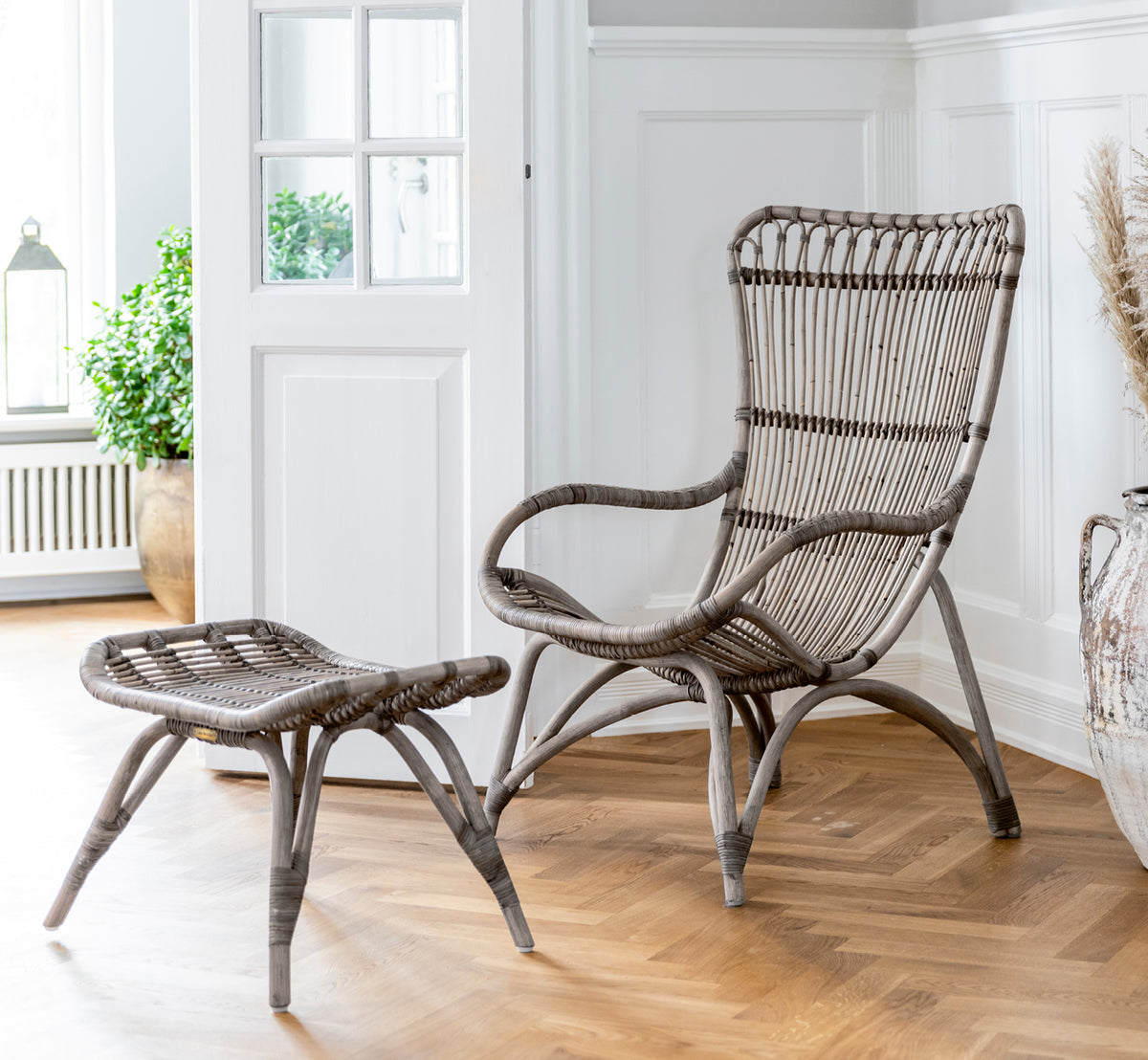 Dies ist ein Lifestyle-Bild von Monet Footstool i taupe rattan. Kombiniert mit Lounge-Sessel in einem dunklen Scheunen-Setting mit Kerze und Glas.