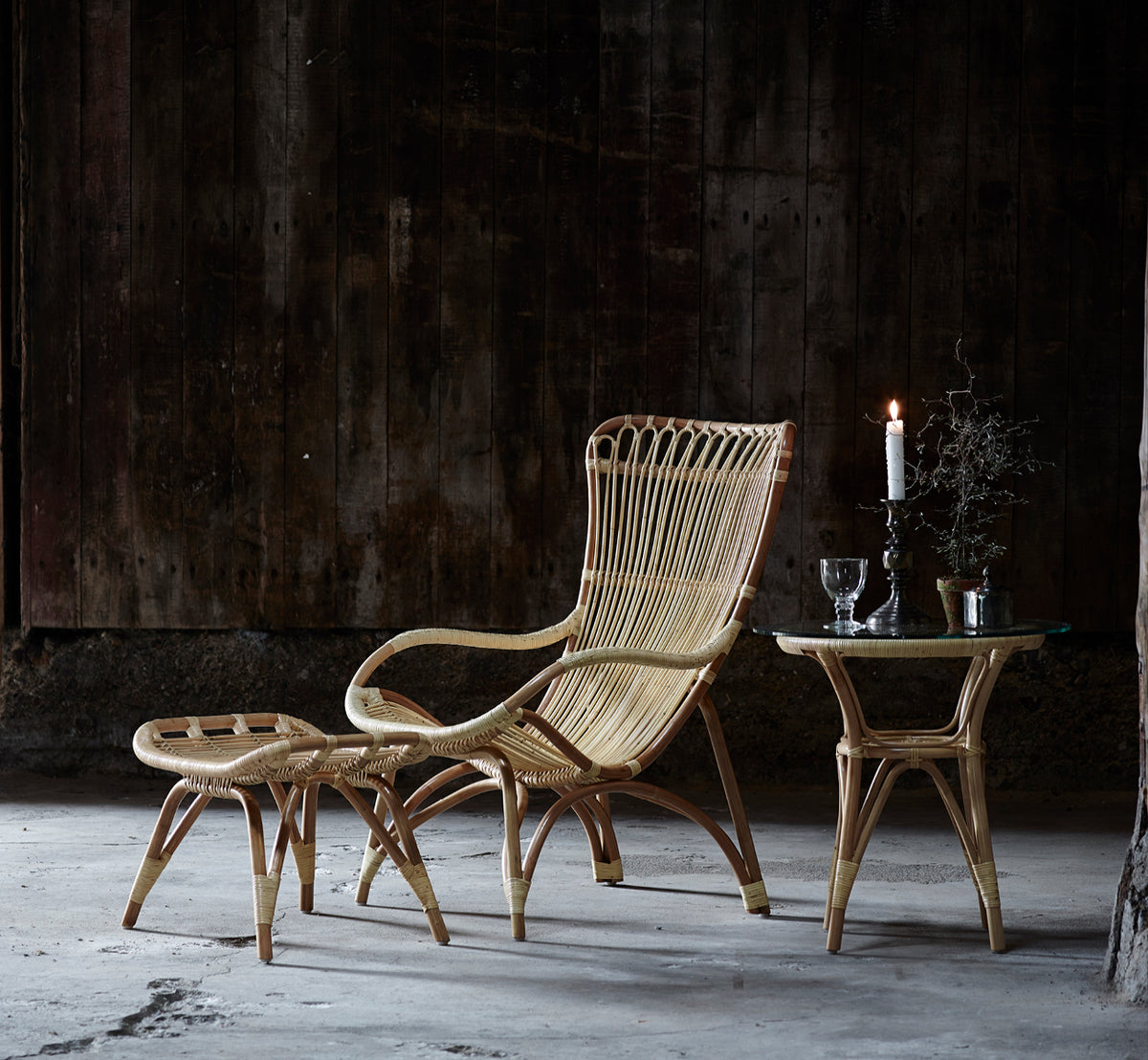 Dies ist ein Lifestyle-Bild von Monet Footstool i natural rattan. Mit Lounge-Sessel in einem dunklen rustikalen Scheunen-Setting mit Glaswaren.