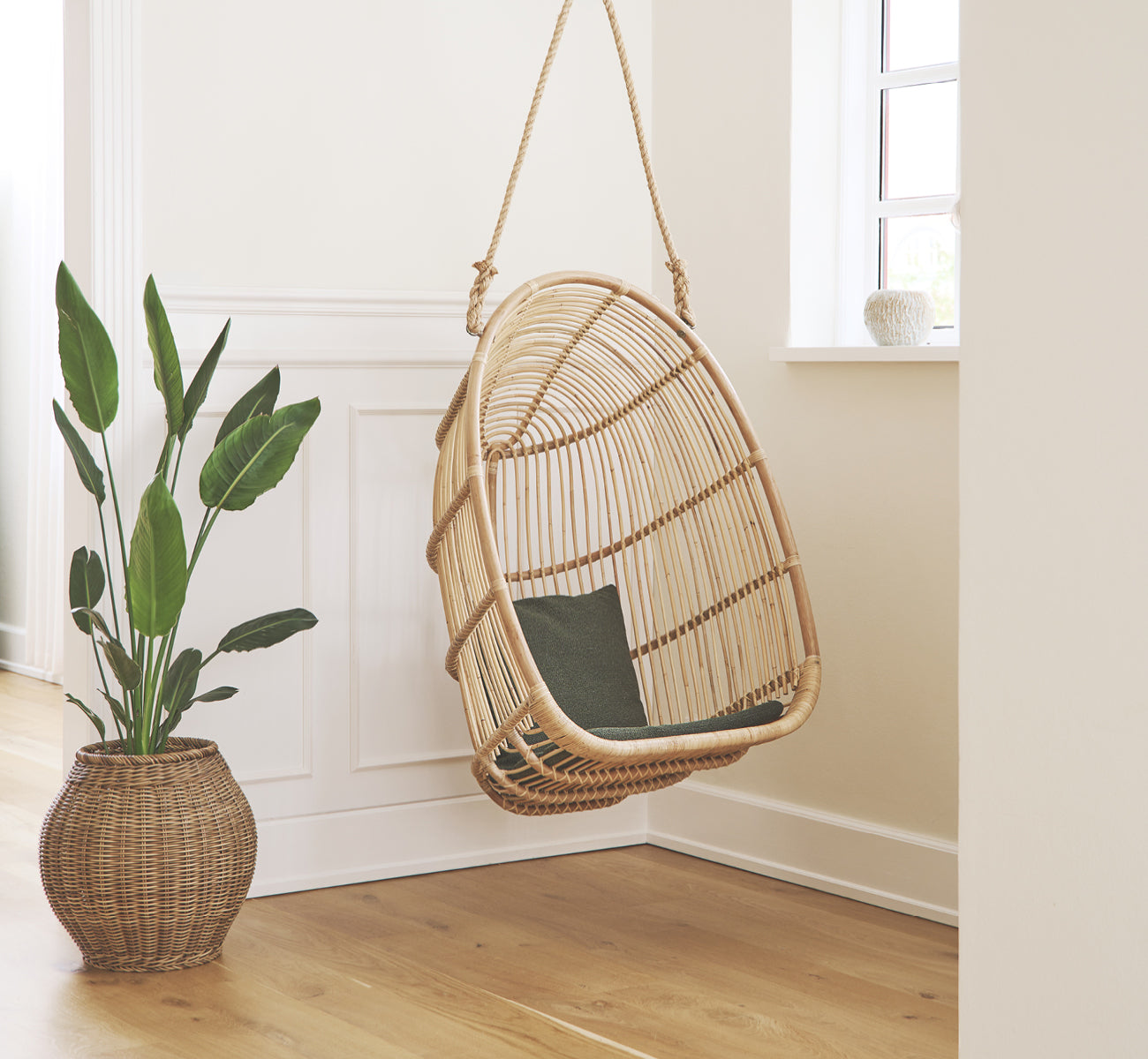 Dies ist ein Lifestyle-Bild von Renoir Hängesessel in natural rattan. In einem hellen vertäfelten Eingang neben einer Blattpflanze hängend.
