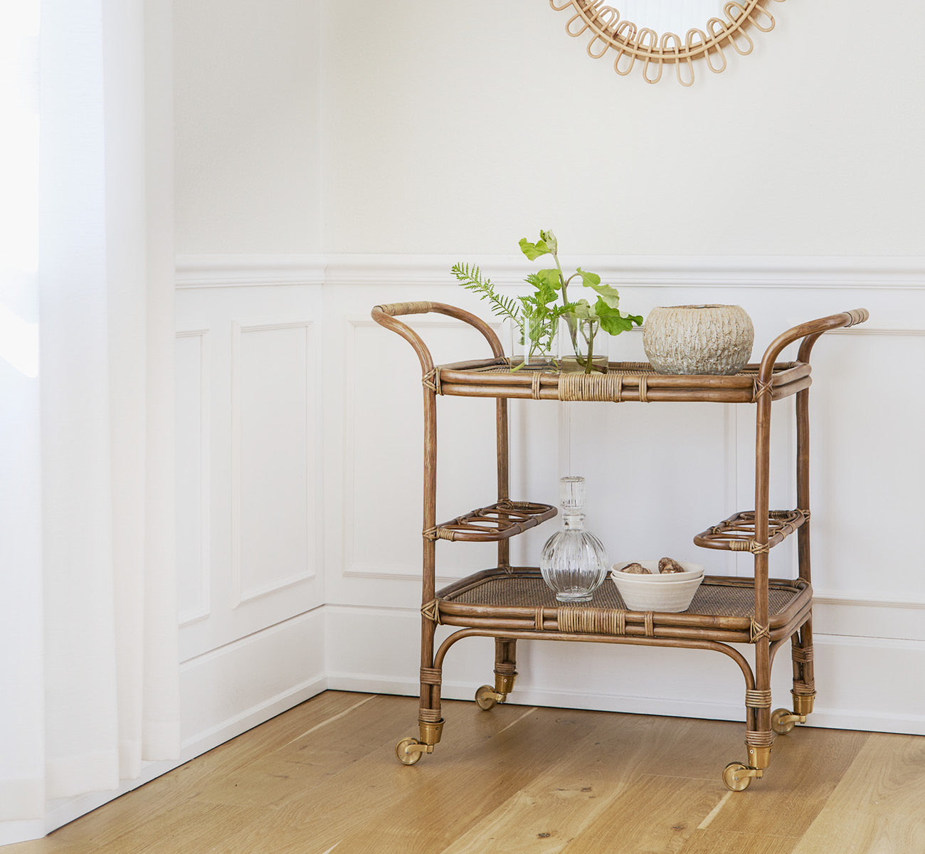Dies ist ein Lifestyle-Bild von Carlo Trolley in antique Rattan, in einem hell-weißen Interieur mit Wandvertäfelung und einem Rattan-Wandspiegel.