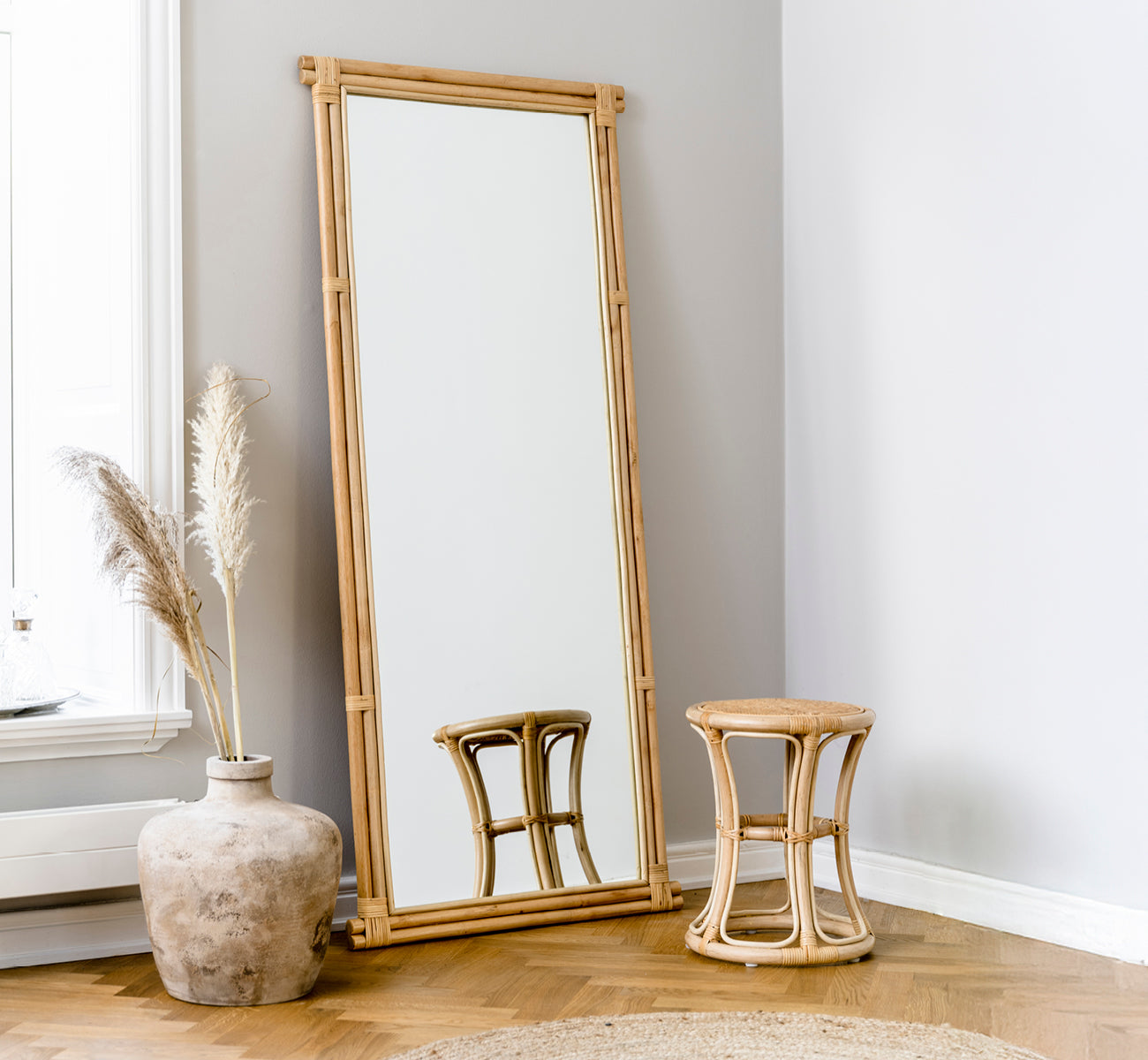 Dies ist ein Lifestyle-Bild von Rasmus Mirror 172 cm in natural rattan. An eine graue Wand neben einem Hocker und getrocknetem Pampasgras gelehnt.