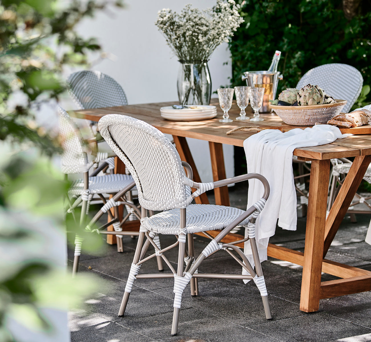 Dies ist ein Lifestyle-Bild von Isabell in taupe aluminium frame Grey w white dot ArtFibre, auf einer üppigen Gartenterrasse mit Steinboden und grün.