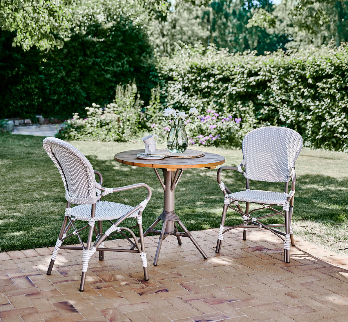 Dies ist ein Lifestyle-Bild von Isabell in taupe aluminium frame Grey w white dot ArtFibre, auf einer üppigen Gartenterrasse mit Steinboden und grün.