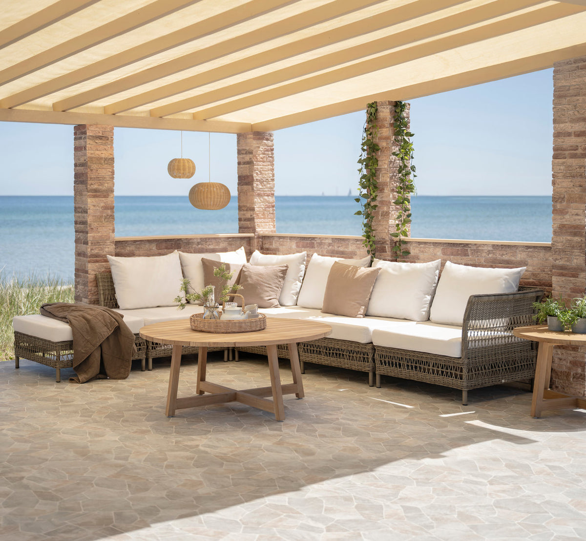 Dies ist ein Lifestyle-Bild von Mona Außen-Modulsofa-Pouf Module in antique grey artfibre aluminium. Unter einer Pergola am Meer.