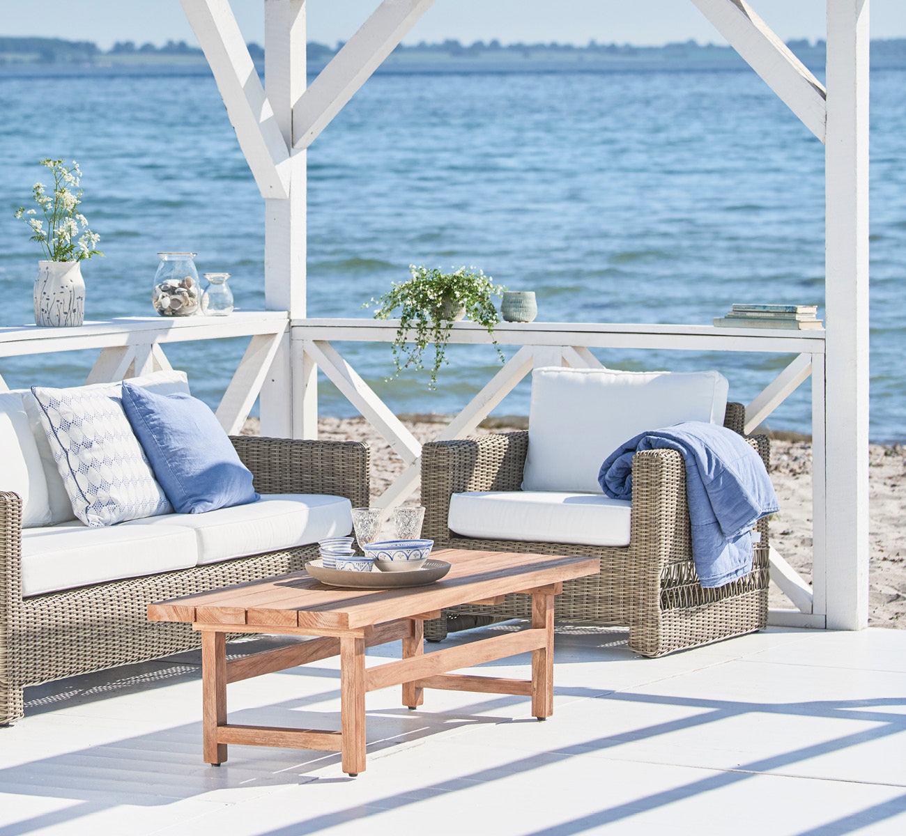 Dies ist ein Lifestyle-Bild von Carrie Lounge-Sessel in antique Grey ArtFibre Aluminium, auf einer sonnendurchfluteten Küstenterrasse mit Meerblick.