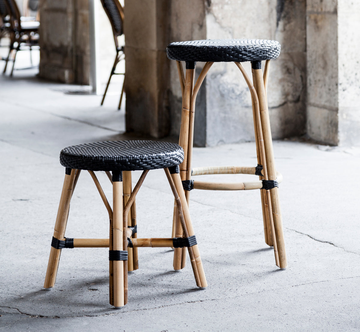 Dies ist ein Lifestyle-Bild von Simone Hocker in black ArtFibre Skin on Rattan frame. An Steinsäulen in einem Café-Ambiente platziert.