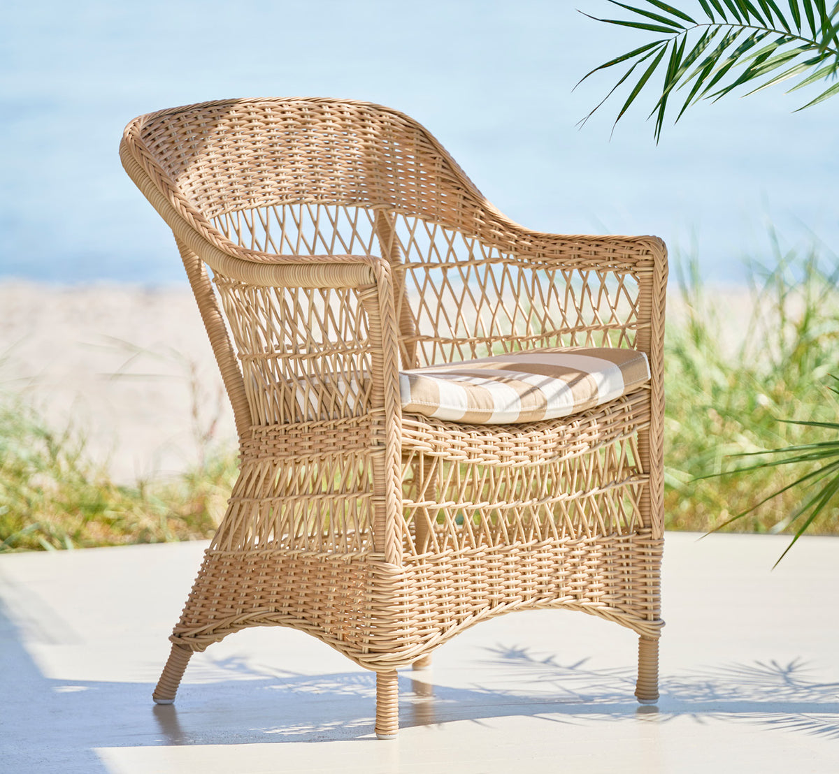Dies ist ein Lifestyle-Bild von Charlot Stuhl in natural ArtFibre Aluminium, auf einer sonnendurchfluteten Küstenterrasse mit Meerblick und einer.