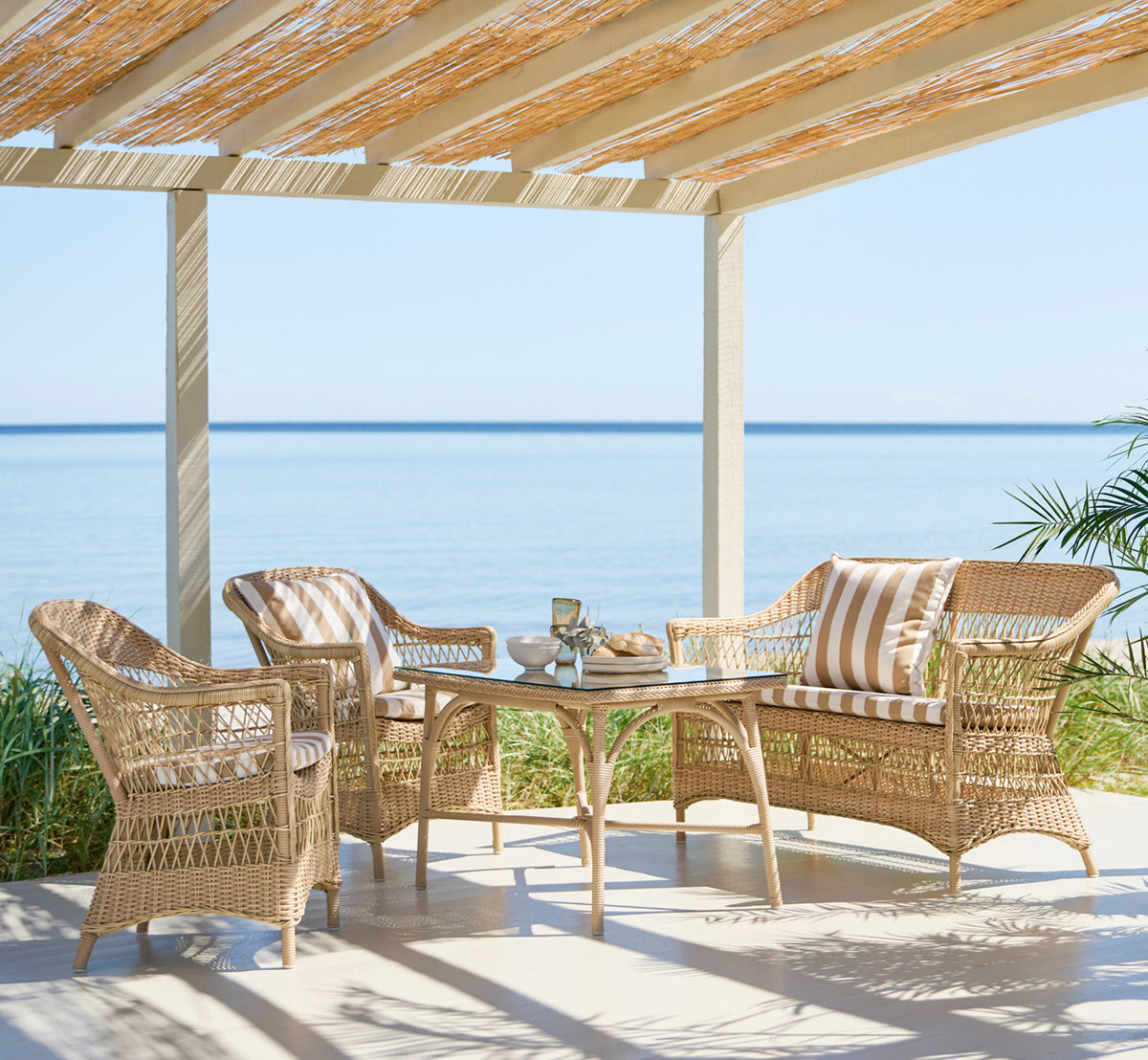 Dies ist ein Lifestyle-Bild von Charlot Stuhl in natural ArtFibre Aluminium, auf einer sonnendurchfluteten Küstenterrasse mit Meerblick und einer.