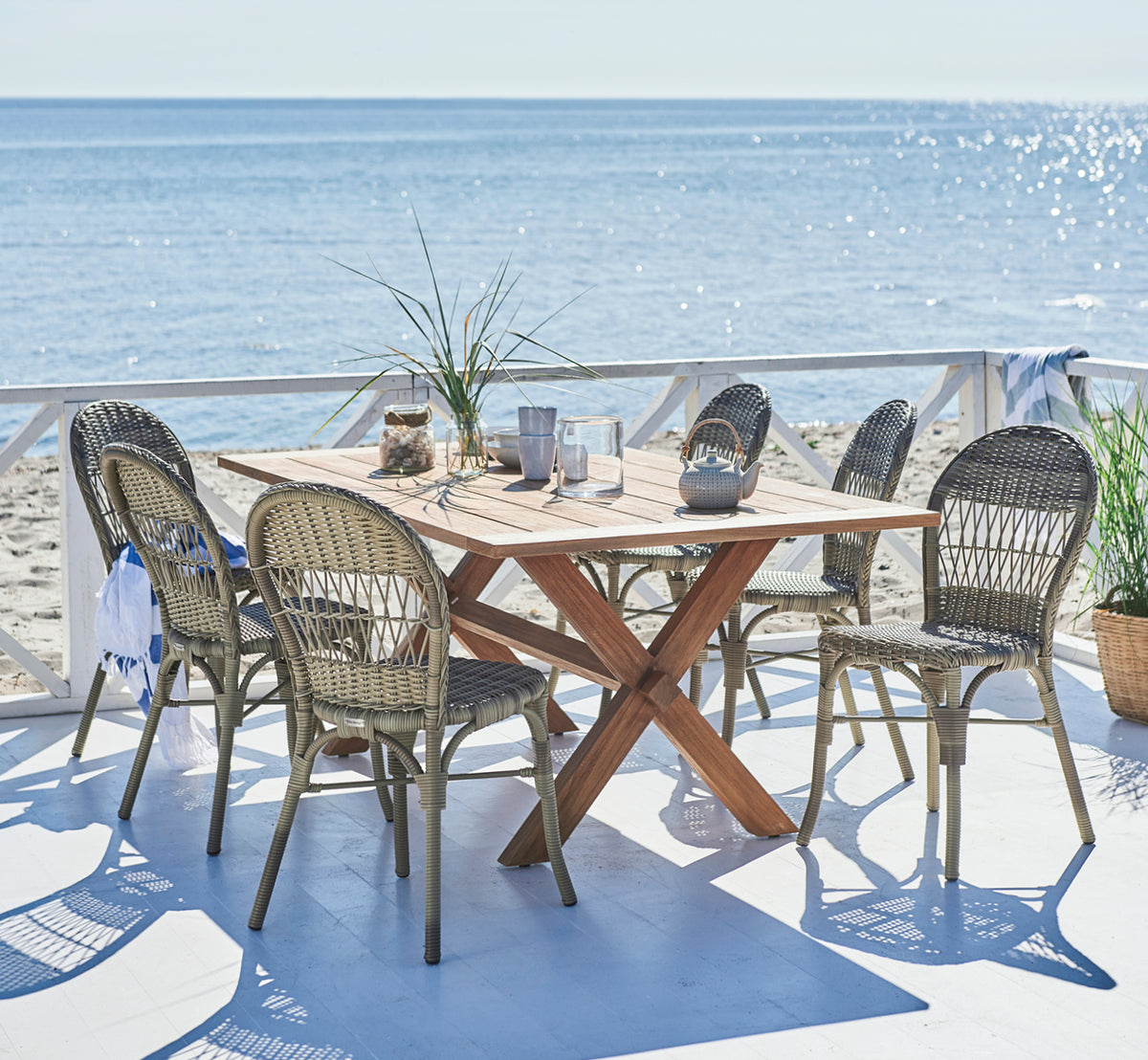 Dies ist ein Lifestyle-Bild von Ofelia Exterior Chair i antique grey artfibre aluminium. Auf einem Stranddeck mit Meerblick und Teekanne.