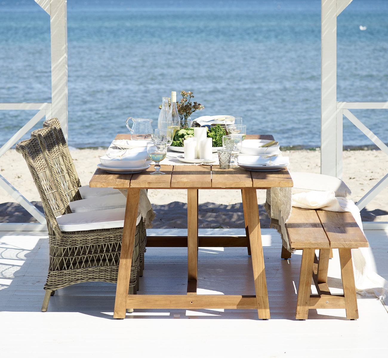 Dies ist ein Lifestyle-Bild von Marie Exterior Side Chair i antique grey artfibre aluminium. An einem Teaktisch in einem Pavillon am Meer.