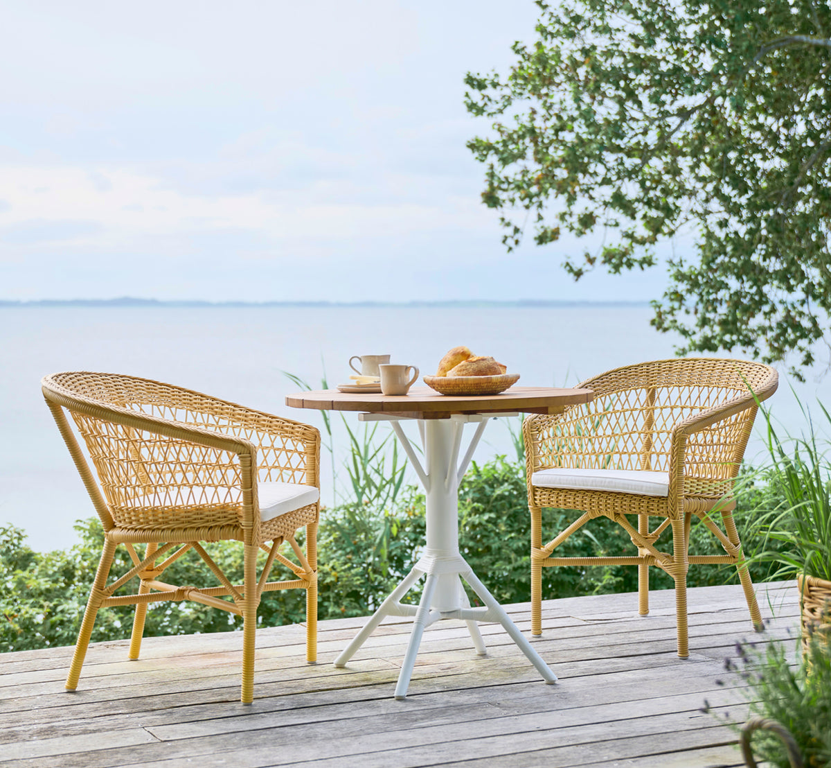 Dies ist ein Lifestyle-Bild von Emma Außen-Esszimmerstuhl in natural ArtFibre Aluminium, auf einer hellen Küstenterrasse mit Schilfdach und Meerblick.