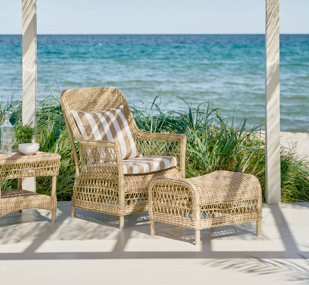 Dies ist ein Lifestyle-Bild von Dawn Fußhocker in natural ArtFibre Aluminium, auf einer Küstenterrasse mit Meerblick, Strandgräsern und weißen Säulen.