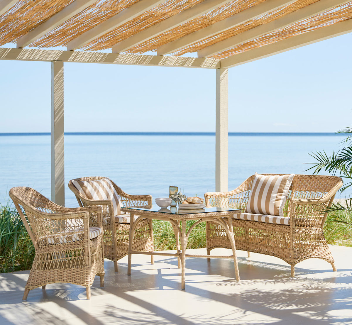 Dies ist ein Lifestyle-Bild von Charlot 2-Sitzer-Sofa in natural ArtFibre Aluminium, auf einer Küstenterrasse mit Schilfdach, Meerblick und.