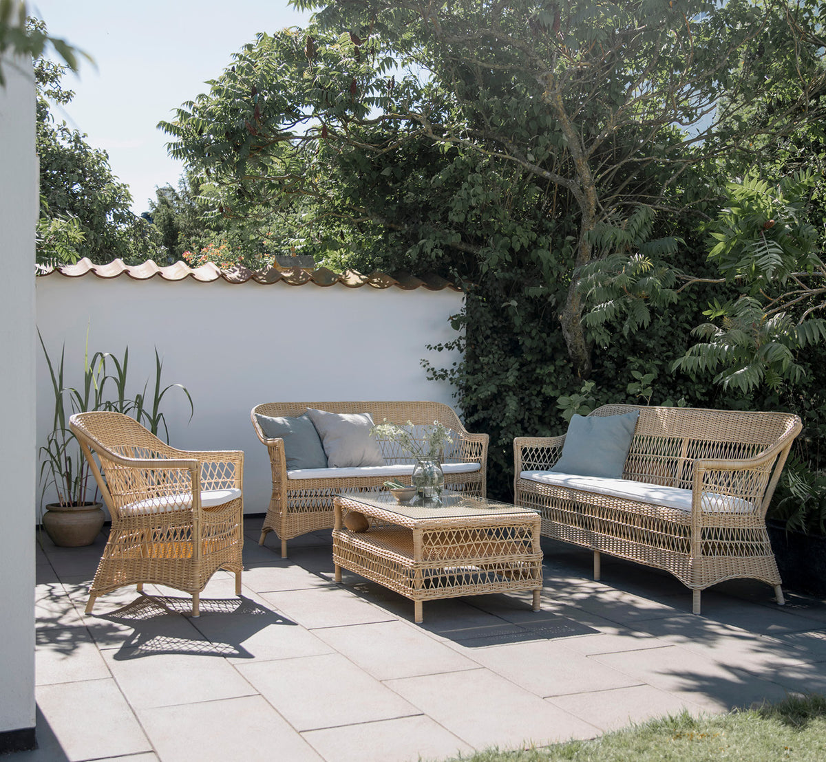 Dies ist ein Lifestyle-Bild von Charlot 2-Sitzer-Sofa in natural ArtFibre Aluminium, auf einer Küstenterrasse mit Schilfdach, Meerblick und.